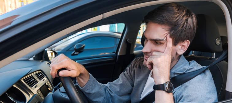 Annoyed tired-looking young man sitting in driver's seat of car with arm on the wheel.