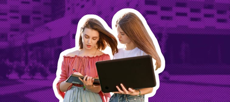 Two college students are standing near the university, looking at a computer, making notes in a notebook.