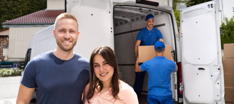 Happy Family Couple And Movers Unload Boxes From Truck.