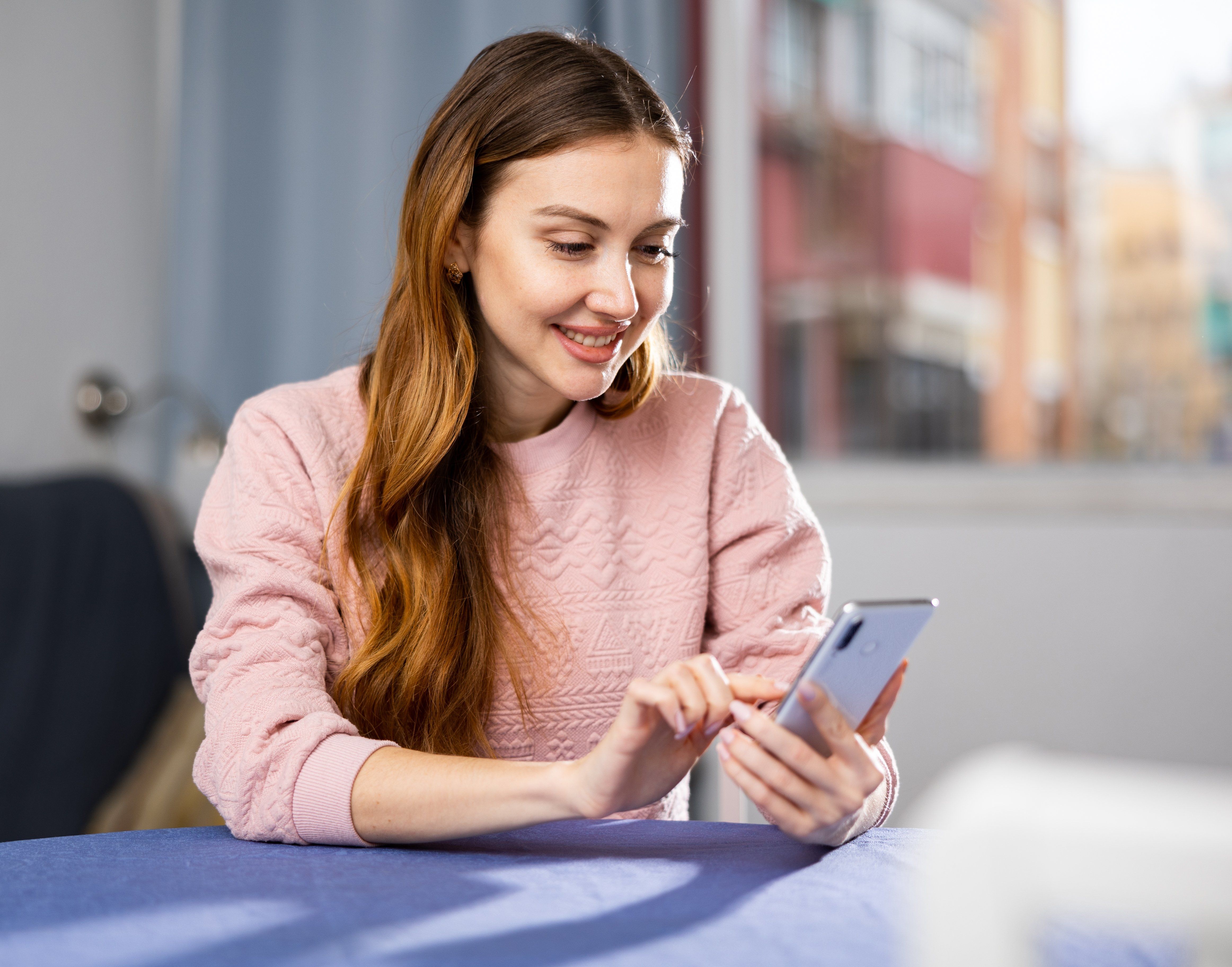 Young smiling woman sitting at a table in an apartment is texting with someone on a mobile phone
