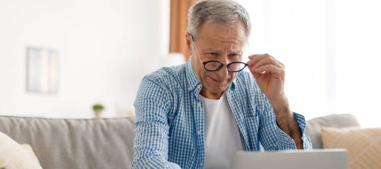 Older man sits on couch looking at computer with puzzled look on his face.