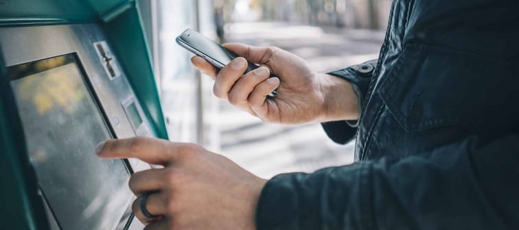 Closeup of male hands using smart phone while typing on ATM, bank machine