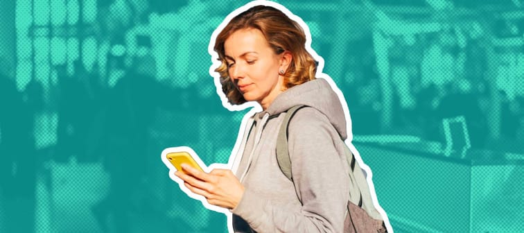 Young woman stands in airport holding handle of suitcase and looking at her phone.