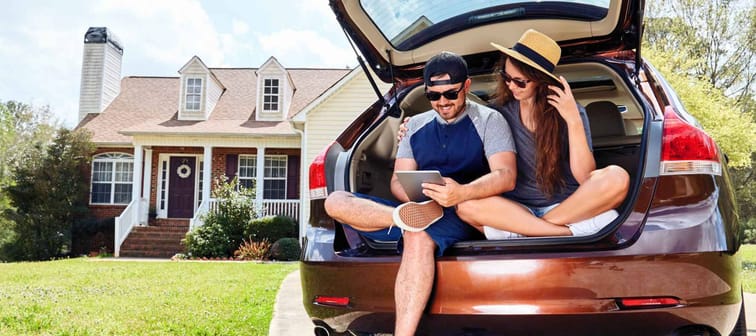 Woman and man sitting in open hatchback of a car, looking at tablet and with their house in the background